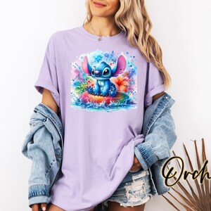Może przedstawiać: Liliowa koszulka z akwarelową ilustracją Stitcha z Lilo & Stitch. Projekt przedstawia Stitcha na kolorowej tratwie z tropikalnymi kwiatami i rozpryskami wody. Koszulka ma kr&oacute;tkie rękawy i luźny kr&oacute;j.