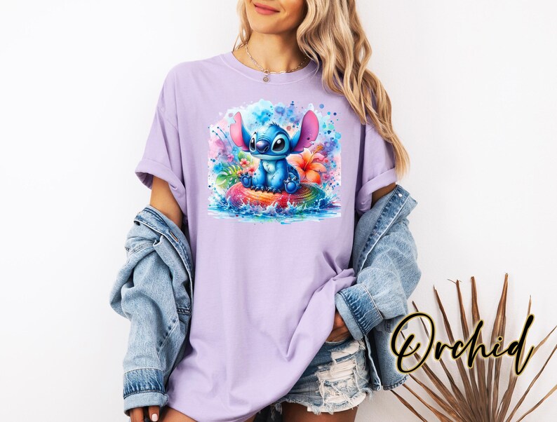 Może przedstawiać: Liliowa koszulka z akwarelową ilustracją Stitcha z Lilo & Stitch. Projekt przedstawia Stitcha na kolorowej tratwie z tropikalnymi kwiatami i rozpryskami wody. Koszulka ma kr&oacute;tkie rękawy i luźny kr&oacute;j.