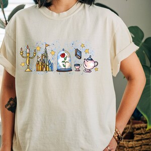 Puede incluir: Una camiseta blanca con un estampado gr&aacute;fico de varios personajes y s&iacute;mbolos de Disney, incluyendo un castillo, una rosa, una tetera y un candelabro. El gr&aacute;fico est&aacute; en un estilo de acuarela con una combinaci&oacute;n de colores azul y amarillo.