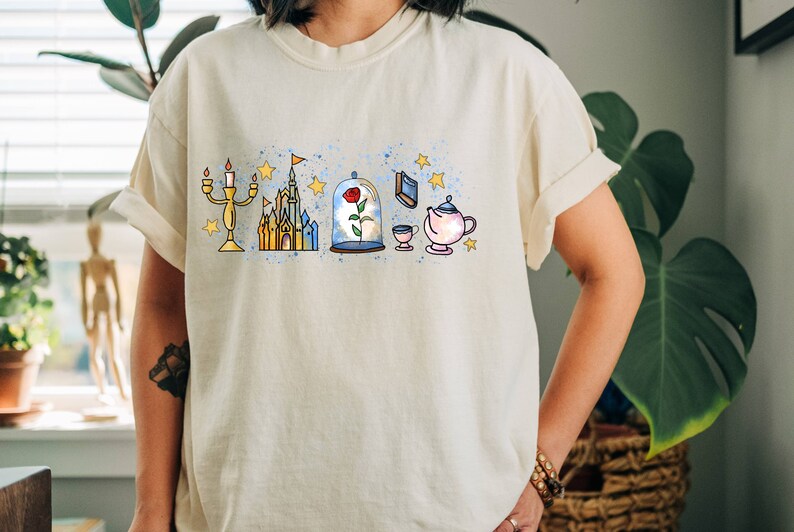 Puede incluir: Una camiseta blanca con un estampado gr&aacute;fico de varios personajes y s&iacute;mbolos de Disney, incluyendo un castillo, una rosa, una tetera y un candelabro. El gr&aacute;fico est&aacute; en un estilo de acuarela con una combinaci&oacute;n de colores azul y amarillo.