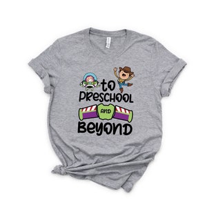 Pode incluir: Camiseta cinza com uma imagem de desenho animado de Woody e Buzz Lightyear de Toy Story. O texto na camiseta diz "to preschool and beyond".