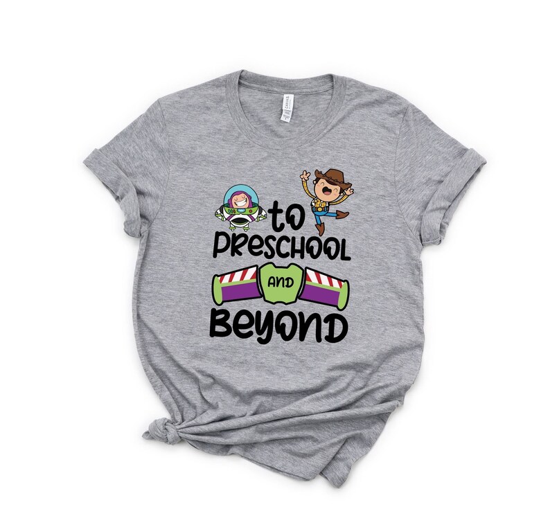 Pode incluir: Camiseta cinza com uma imagem de desenho animado de Woody e Buzz Lightyear de Toy Story. O texto na camiseta diz "to preschool and beyond".