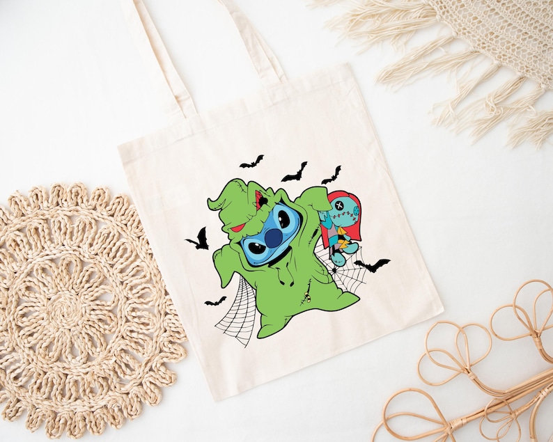 May include: A cream-colored tote bag featuring a Halloween-themed graphic. The design includes Stitch dressed as Oogie Boogie, with Sally from The Nightmare Before Christmas. Black bats and a spider web complete the spooky design.