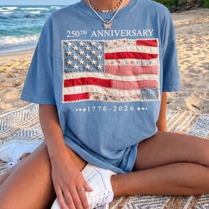 May include: A light blue t-shirt featuring a graphic of an American flag composed of various patterned fabric patches. The text 250TH ANNIVERSARY is printed above the flag, and 1776-2026 is printed below, flanked by three stars on each side.
