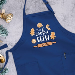 May include: Blue kitchen apron with a white design that says "The Cookie Crew" with gingerbread cookies and a rolling pin. The text "NAME" is on the rolling pin.