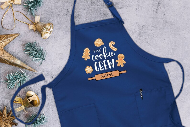 May include: Blue kitchen apron with a white design that says "The Cookie Crew" with gingerbread cookies and a rolling pin. The text "NAME" is on the rolling pin.
