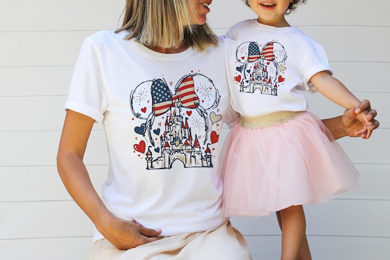 May include: White t-shirts with a patriotic design featuring a castle, Mickey Mouse ears with an American flag, and red and blue hearts. The shirts are worn by a mother and child, with the child wearing a pink tutu.