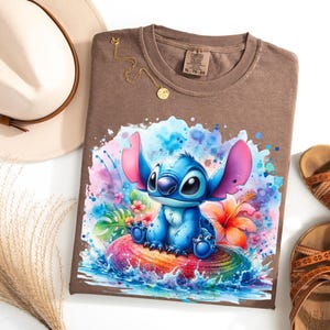 Może przedstawiać: Brązowa koszulka z akwarelową ilustracją postaci Stitch z filmu Lilo & Stitch. Projekt przedstawia Stitcha siedzącego na kolorowej skale z tropikalnymi kwiatami i rozpryskami wody. Na zdjęciu widoczny jest r&oacute;wnież złoty naszyjnik, sandały i kapelusz.