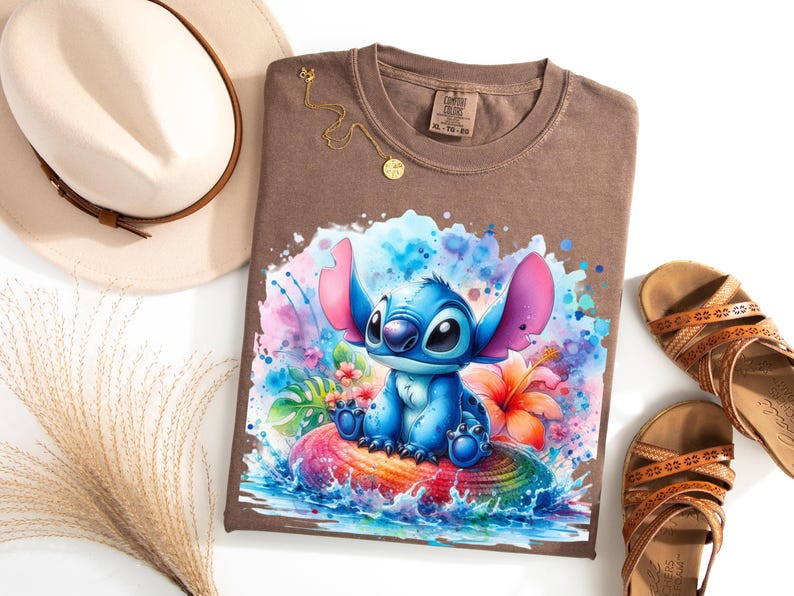 Może przedstawiać: Brązowa koszulka z akwarelową ilustracją postaci Stitch z filmu Lilo & Stitch. Projekt przedstawia Stitcha siedzącego na kolorowej skale z tropikalnymi kwiatami i rozpryskami wody. Na zdjęciu widoczny jest r&oacute;wnież złoty naszyjnik, sandały i kapelusz.