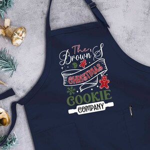Puede incluir: Un delantal azul marino con un bolsillo blanco y un dise&ntilde;o festivo que dice "The Brown's Christmas Cookie Company" con un hombre de jengibre y un rodillo.