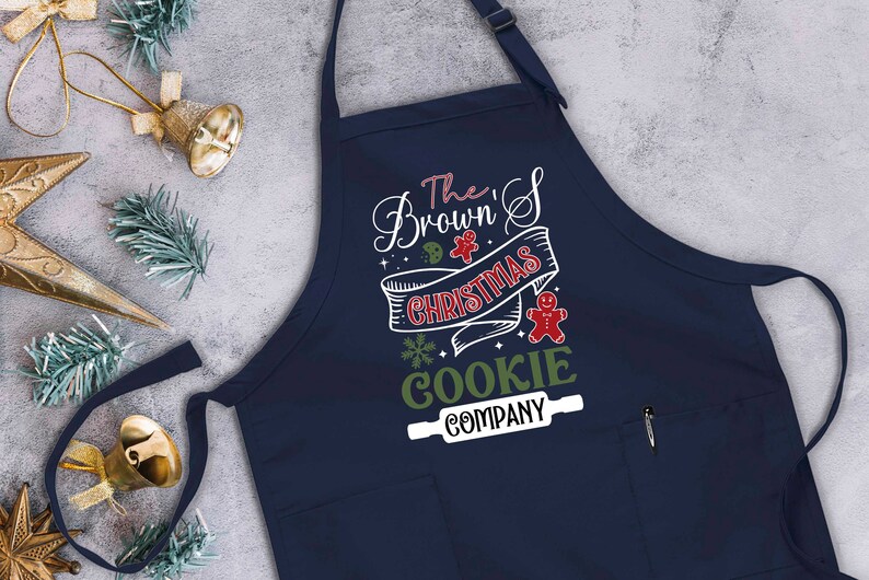 Puede incluir: Un delantal azul marino con un bolsillo blanco y un dise&ntilde;o festivo que dice "The Brown's Christmas Cookie Company" con un hombre de jengibre y un rodillo.