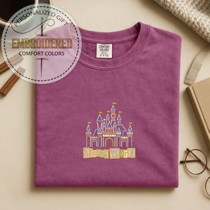 May include: A plum-colored t-shirt with an embroidered design of a castle and the words "Disney World" in yellow. The shirt has a crew neck and short sleeves. The words "Embroidered Comfort Colors" are in a circular design in the upper left corner.