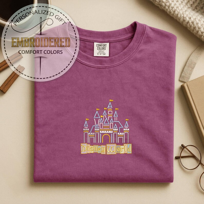 May include: A plum-colored t-shirt with an embroidered design of a castle and the words "Disney World" in yellow. The shirt has a crew neck and short sleeves. The words "Embroidered Comfort Colors" are in a circular design in the upper left corner.
