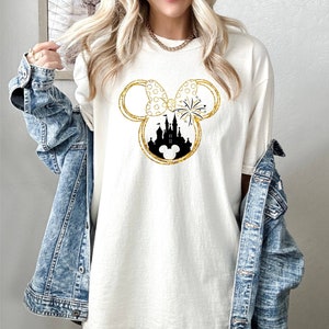 May include: A white t-shirt with a gold glitter design of Mickey Mouse ears with a silhouette of Cinderella's castle inside the ears. The bow on the ears has white polka dots.
