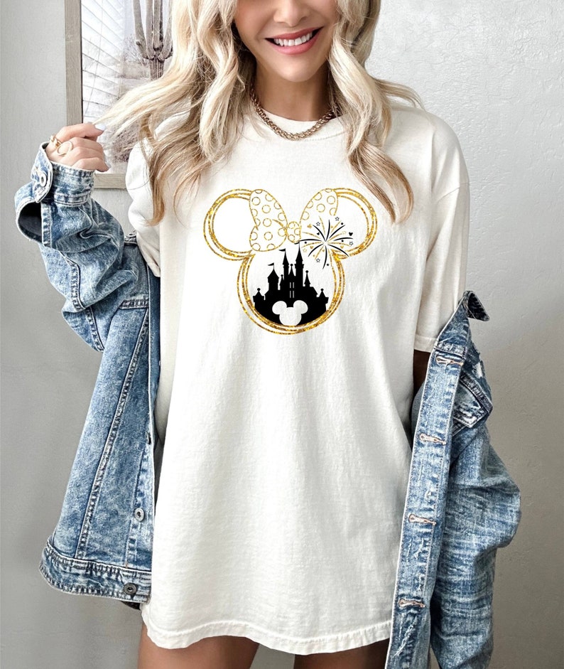 May include: A white t-shirt with a gold glitter design of Mickey Mouse ears with a silhouette of Cinderella's castle inside the ears. The bow on the ears has white polka dots.