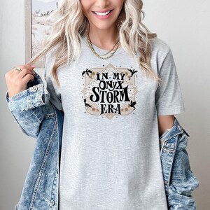 May include: A light gray t-shirt with the text "IN MY ONYX STORM ERA" in a decorative design. The design includes black silhouettes and a tan border. The shirt is made of a soft material.