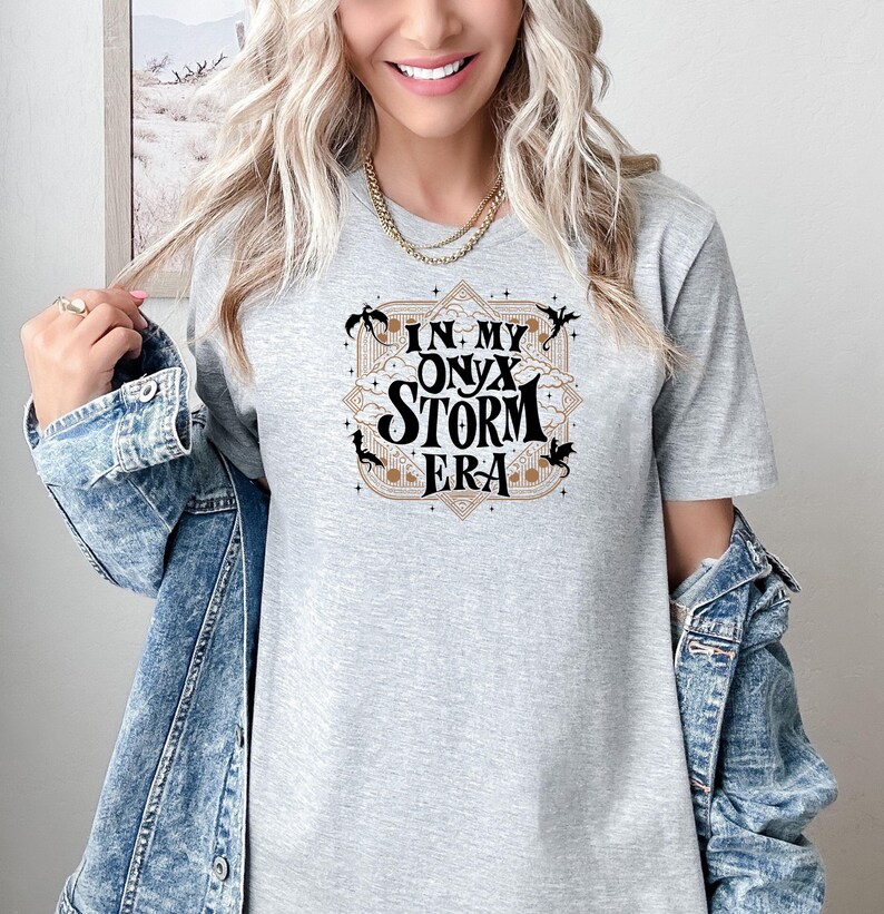 May include: A light gray t-shirt with the text "IN MY ONYX STORM ERA" in a decorative design. The design includes black silhouettes and a tan border. The shirt is made of a soft material.