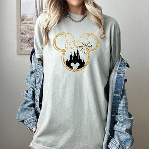May include: A light green t-shirt with a gold glitter Mickey Mouse silhouette featuring a black silhouette of Cinderella's castle and fireworks inside the ears.