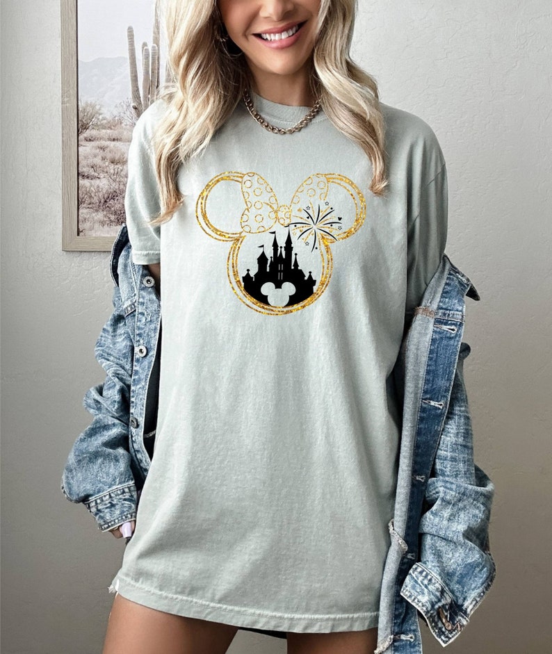 May include: A light green t-shirt with a gold glitter Mickey Mouse silhouette featuring a black silhouette of Cinderella's castle and fireworks inside the ears.