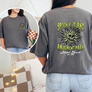 May include: Dark gray t-shirt with a graphic on the back that reads "Spooky Island Amusement Park Island Resort Est. 2002." The graphic features a skull with a sunburst design in green and yellow. The front of the shirt has a smaller version of the graphic.