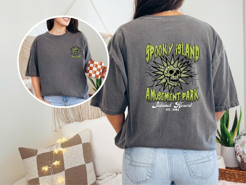 May include: Dark gray t-shirt with a graphic on the back that reads "Spooky Island Amusement Park Island Resort Est. 2002." The graphic features a skull with a sunburst design in green and yellow. The front of the shirt has a smaller version of the graphic.