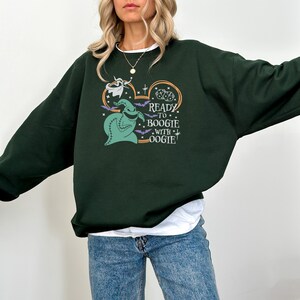 Pu&ograve; includere: Felpa verde scuro con una grafica di Oogie Boogie da The Nightmare Before Christmas. Il design include il testo "Ready to Boogie with Oogie", pipistrelli e stelle. La felpa &egrave; uno stile casual e confortevole.