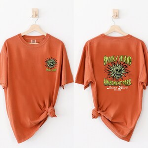 May include: Burnt orange t-shirts with a spooky theme. One shirt features a small green skull and sunburst design. The other shirt has a larger design on the back with the text "Spooky Island Amusement Park Island Resort Est. 2002".