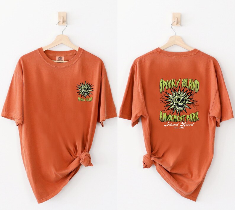 May include: Burnt orange t-shirts with a spooky theme. One shirt features a small green skull and sunburst design. The other shirt has a larger design on the back with the text "Spooky Island Amusement Park Island Resort Est. 2002".