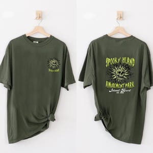 May include: Olive green t-shirts with a graphic design. One shirt features a skull and the words "Spooky Island" in neon green. The other shirt has the same design on the front and the words "Spooky Island Amusement Park" on the back.