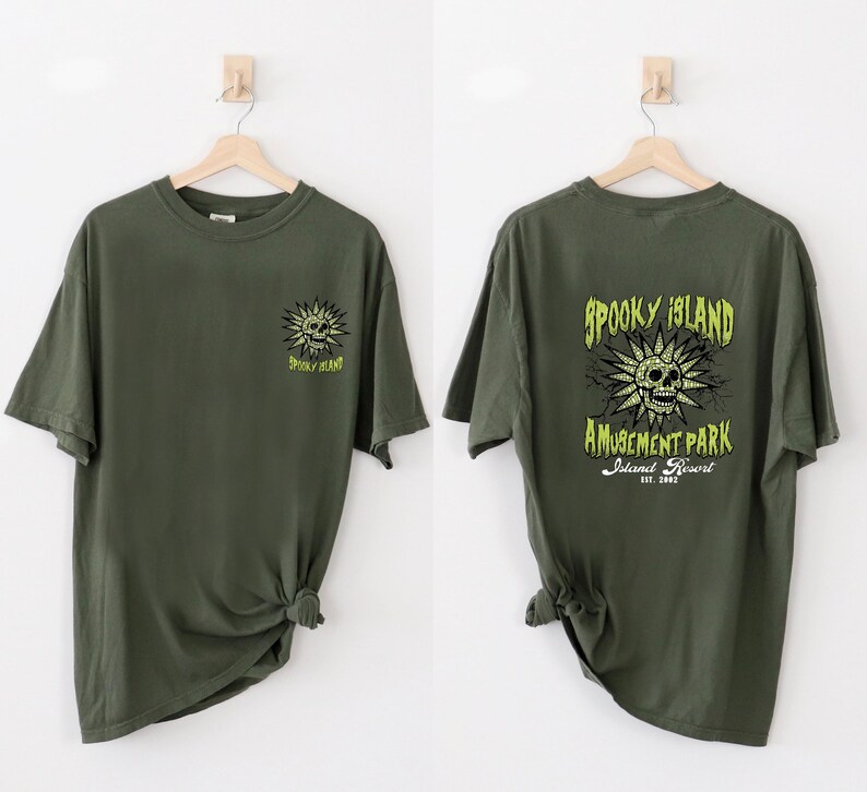 May include: Olive green t-shirts with a graphic design. One shirt features a skull and the words "Spooky Island" in neon green. The other shirt has the same design on the front and the words "Spooky Island Amusement Park" on the back.
