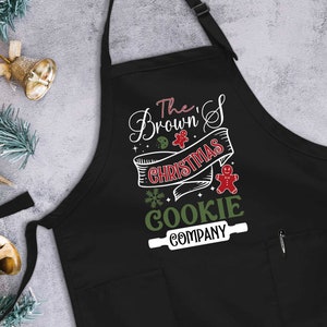 Puede incluir: Delantal negro con tem&aacute;tica navide&ntilde;a con texto blanco que dice "The Brown's Christmas Cookie Company" con una galleta verde y blanca y un hombre de jengibre rojo.