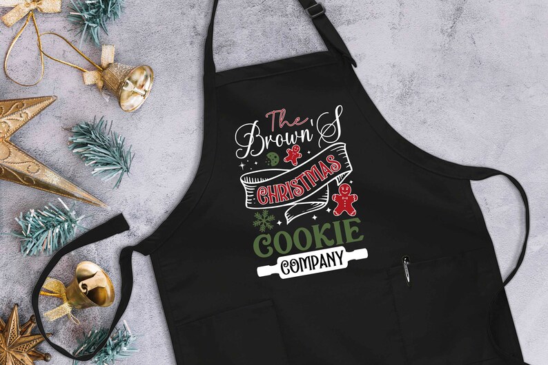 Puede incluir: Delantal negro con tem&aacute;tica navide&ntilde;a con texto blanco que dice "The Brown's Christmas Cookie Company" con una galleta verde y blanca y un hombre de jengibre rojo.