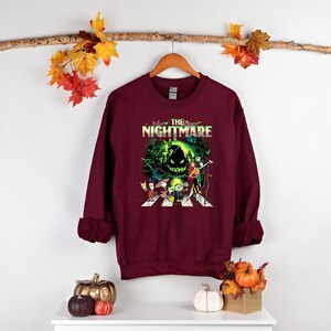 May include: Burgundy sweatshirt featuring a graphic from "The Nightmare Before Christmas." The design includes characters crossing a crosswalk with the film's title above. The sweatshirt is hanging on a wooden hanger.