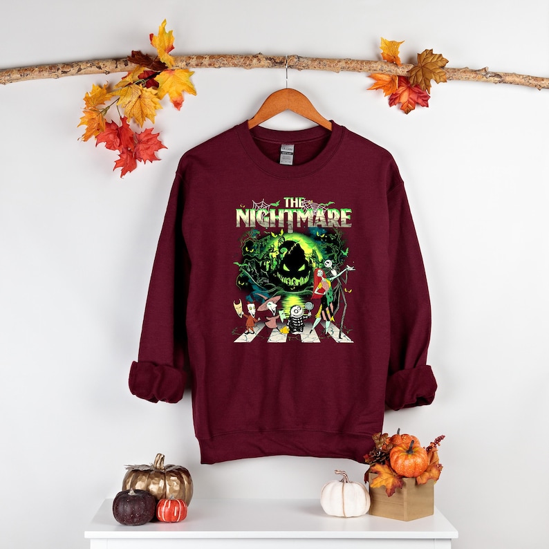 May include: Burgundy sweatshirt featuring a graphic from "The Nightmare Before Christmas." The design includes characters crossing a crosswalk with the film's title above. The sweatshirt is hanging on a wooden hanger.