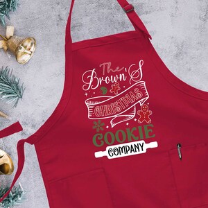 Puede incluir: Delantal rojo con tem&aacute;tica navide&ntilde;a con un dise&ntilde;o blanco que dice "The Brown's Christmas Cookie Company" con un hombre de jengibre y un rodillo.