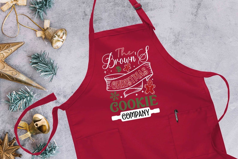 Puede incluir: Delantal rojo con tem&aacute;tica navide&ntilde;a con un dise&ntilde;o blanco que dice "The Brown's Christmas Cookie Company" con un hombre de jengibre y un rodillo.