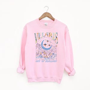 May include: A light pink sweatshirt with the word "VELARIS" in colorful lettering above a crescent moon and stars design. Below the design is the text "CITY OF STARLIGHT". The sweatshirt is hanging on a wooden hanger.