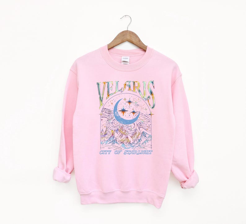 May include: A light pink sweatshirt with the word "VELARIS" in colorful lettering above a crescent moon and stars design. Below the design is the text "CITY OF STARLIGHT". The sweatshirt is hanging on a wooden hanger.