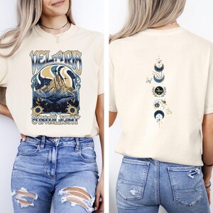 May include: Cream-colored t-shirt featuring a graphic design on the front with the text "VELARIS CITY OF STARLIGHT" above a mountain scene with sunflowers. The back of the shirt displays a moon phase design with butterflies.
