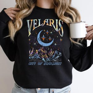 May include: Black sweatshirt with a celestial design featuring a crescent moon, stars, and mountains. The text "VELARIS CITY OF STARLIGHT" is arched above the moon. A white mug is held in the hand.