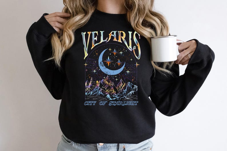May include: Black sweatshirt with a celestial design featuring a crescent moon, stars, and mountains. The text "VELARIS CITY OF STARLIGHT" is arched above the moon. A white mug is held in the hand.