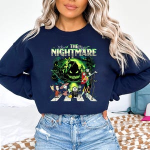 May include: Navy blue sweatshirt featuring a graphic from "The Nightmare Before Christmas." The design includes the text "The Nightmare" above a scene with Jack Skellington, Sally, and other characters crossing a crosswalk in front of a spooky green background.