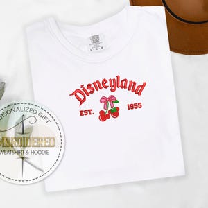 Op de afbeelding: Wit T-shirt met rode geborduurde tekst die "Disneyland Est. 1955" zegt met een rode kers en een roze strik.