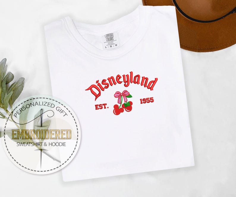 Op de afbeelding: Wit T-shirt met rode geborduurde tekst die "Disneyland Est. 1955" zegt met een rode kers en een roze strik.
