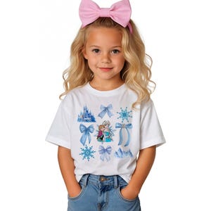 May include: White t-shirt featuring a design with blue bows, snowflakes, and a castle. The shirt also has an image of two characters from the movie Frozen. A pink bow headband is worn by the person in the image.
