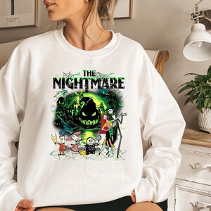 May include: White sweatshirt featuring a graphic from "The Nightmare Before Christmas." The design includes the text "THE NIGHTMARE" above a scene with Jack Skellington, Sally, and other characters in a spooky, green-toned setting.