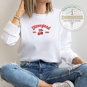 May include: White sweatshirt with a red embroidered logo that says "Disneyland Est. 1955" with a red bow and cherries.