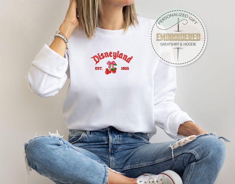 May include: White sweatshirt with a red embroidered logo that says "Disneyland Est. 1955" with a red bow and cherries.