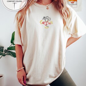 May include: A white t-shirt with a simple line drawing of Mickey Mouse embroidered on the front. The embroidery is in red, black, and yellow thread.