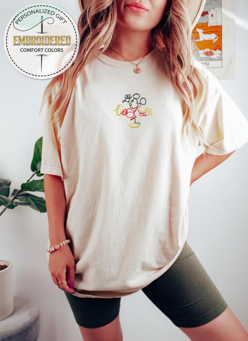 May include: A white t-shirt with a simple line drawing of Mickey Mouse embroidered on the front. The embroidery is in red, black, and yellow thread.